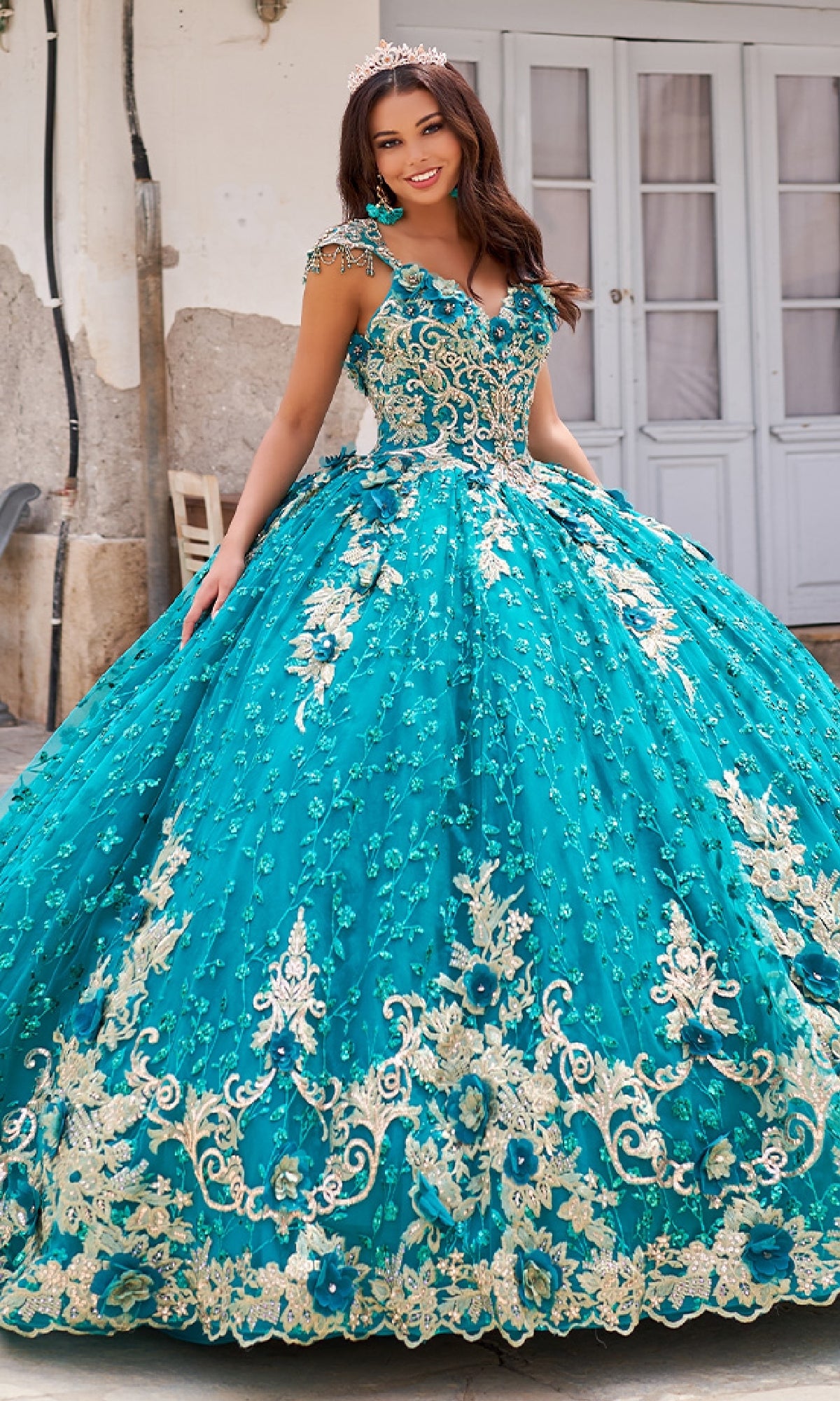 A woman in the Quinceanera Dress PR30088 By Princesa, featuring teal and gold lace embroidery, a full skirt, and a tiara, smiles outdoors in front of white double doors, perfectly showcasing this stunning gown.